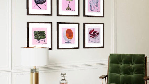 A styled interior scene showing a wall gallery of nine framed iconic design art prints with pink backgrounds and artist signature (Freddie Farrow), above a round glass-top coffee table and next to a green tufted armchair.